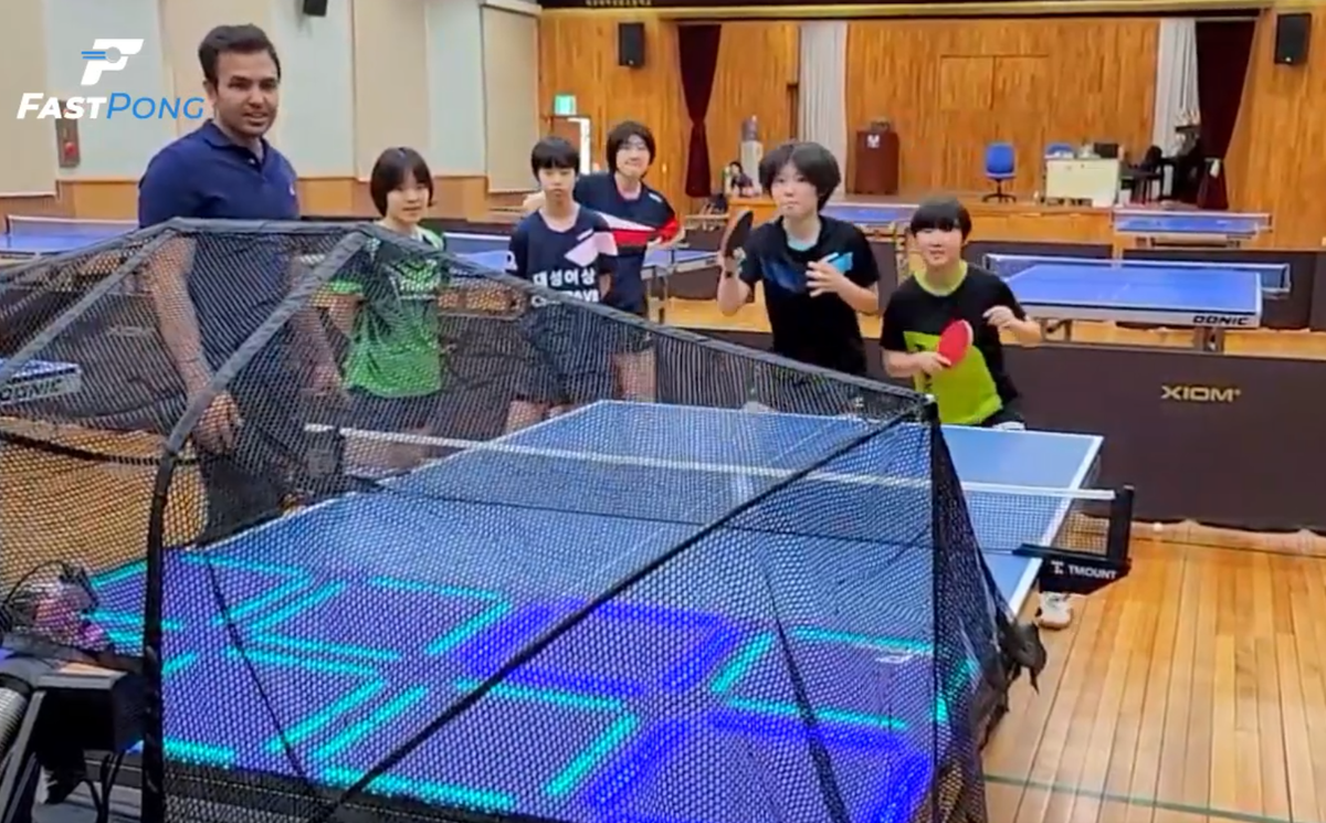 In a gymnasium is full of table tennis tables, two table tennis players are having a competitive game of Green versus Blue while other players watch