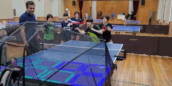In a gymnasium filled with table tennis tables two table tennis players play a game of green versus blue on the Fastpong system while other players watch